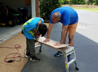 Graham holding down unfinised skateboard while Joseph use power tool to drill a hole in the skateboard