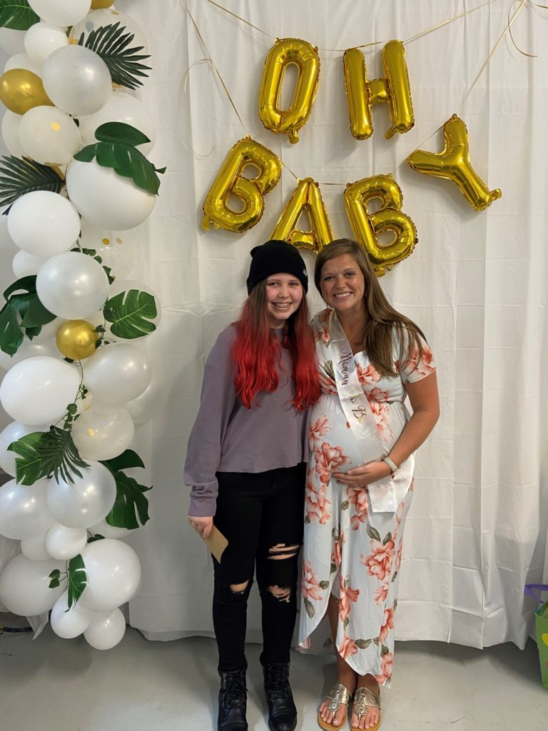 Pregent Nikki and older Kayley, smiling in front of balloons that say "Oh Baby"
