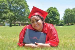 Graduate in red cap and gown holding a diploma lies on their stomach in the green grass looking at the camera