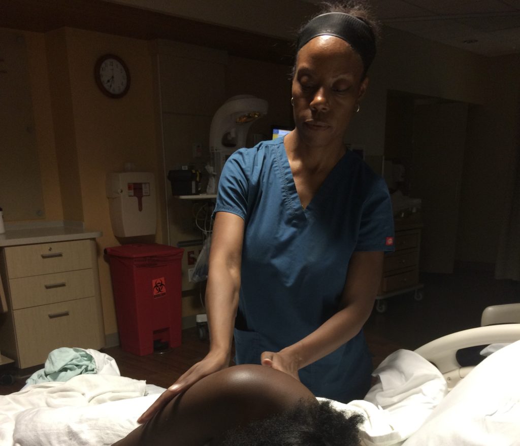 Black female doula performing a massage on a birthing individual in labor in a hospital room.