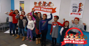 Girls replicating the Rosie Riveter arm and fist in air pose, standing in front of a sign in a classroom